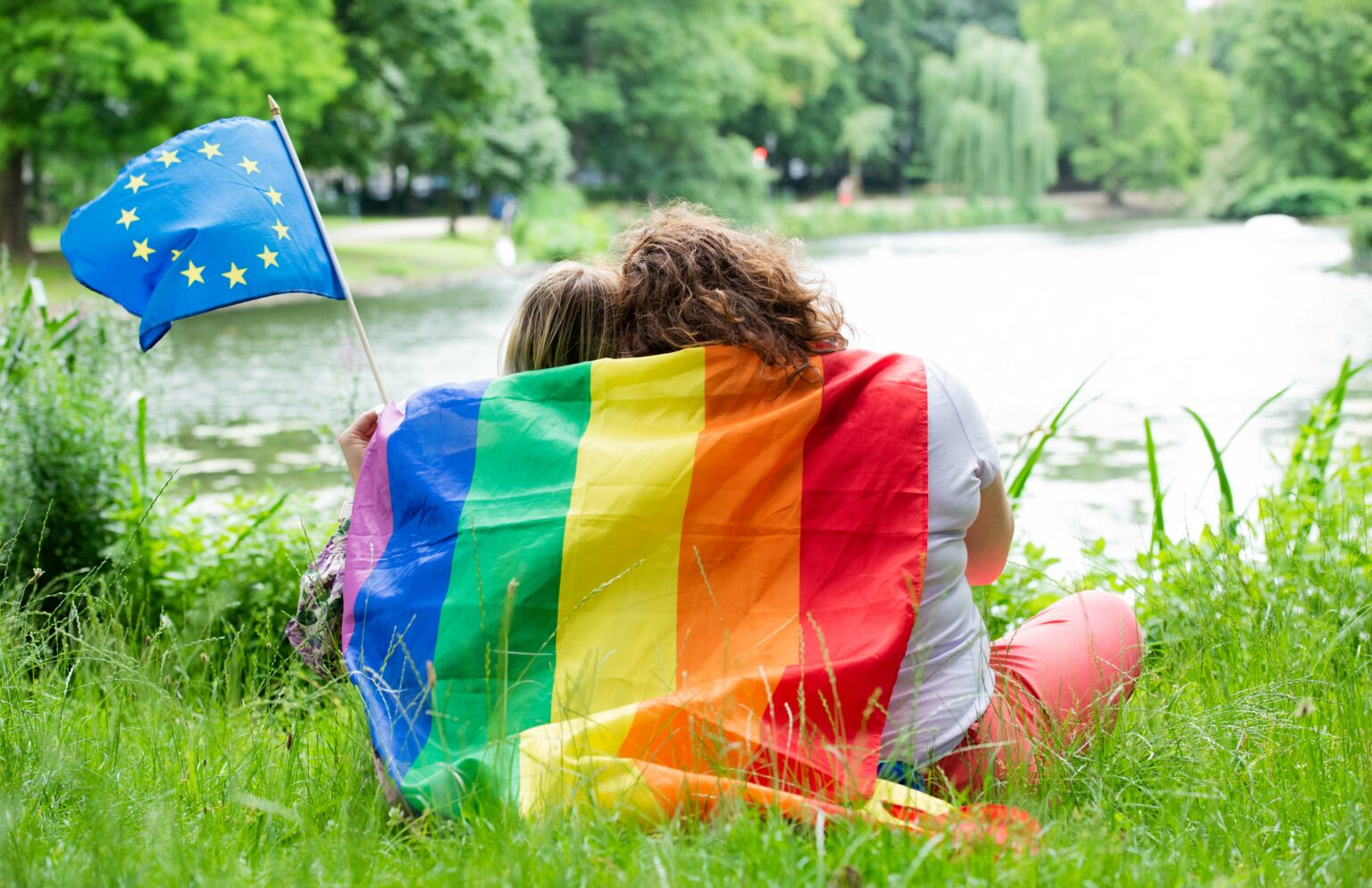 Two people wrapped in the rainbow flag and waving a small EU flag.