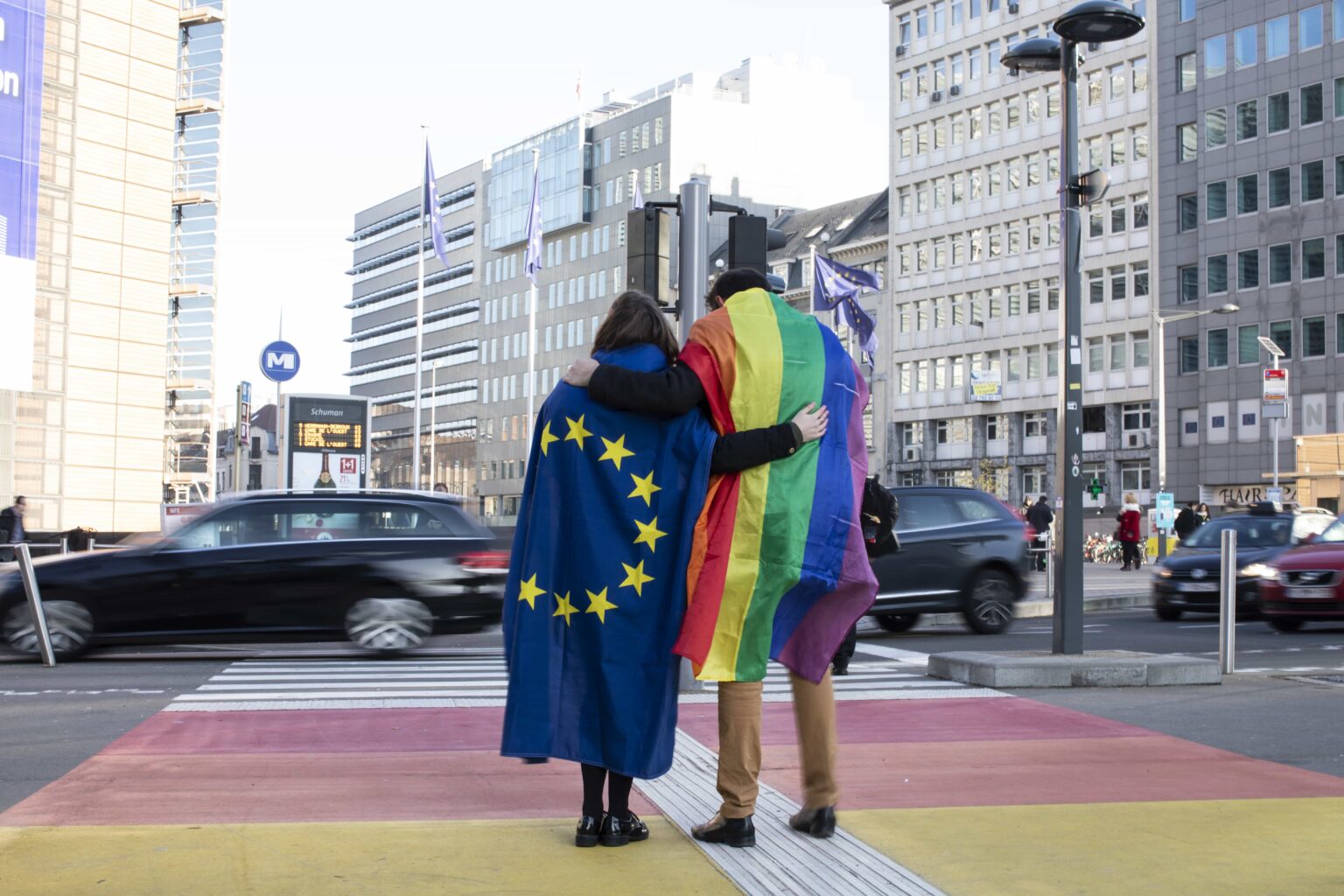 Two people wear EU and rainbow flags on a crosswalk painted in rainbow colors